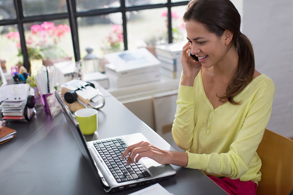 Smiling woman working at laptop and talking on smart phone in office